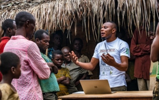 person talking to a group of people with a computer Infront of him and a object in his hand